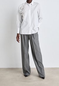 Camicia bianca con bottoni, con un dettaglio floreale plissettato sulla spalla sinistra, abbinata a pantaloni larghi a quadri grigi e scarpe nere con punta.
