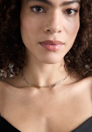 Metal necklace with alternating silver and gold links, featuring elongated shapes and a mix of textures. Worn around the neckline.