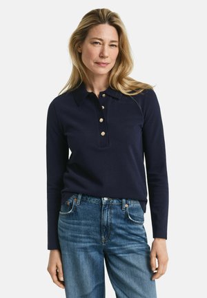 Vrouw met blond haar, gekleed in een marineblauwe lange mouwen polo shirt met gouden knopen en blauwe denim jeans, staand tegen een witte achtergrond.