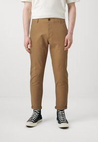 Brown cotton chinos with a straight cut, featuring a single button closure. Paired with black high-top sneakers with white soles.