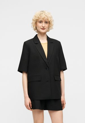 Jeune personne aux cheveux blonds bouclés et courts portant un blazer noir à manches courtes et un short noir assorti par-dessus un haut jaune.