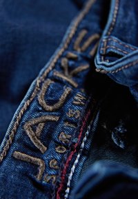 Ciemnoniebieski dżins z złotym haftem z napisem "JACKS." Wyróżniają się widoczne szwy w odcieniach beżu i czerwieni oraz teksturowana powierzchnia materiału.