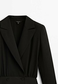 Blazer noir cintré avec col à revers cranté, manches longues et détail de double boutonnage, présenté sur fond blanc.