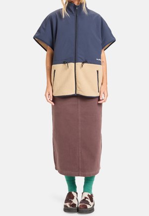 Persoon draagt een marineblauwe en beige gilet met ritssluiting, een bruine midirok, groene sokken en bruine-witte slip-on schoenen met patroon.