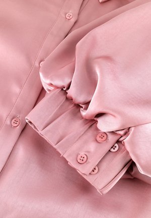 Zachte roze satijnen blouse met lange geplooide mouwen en met knopen gestikte manchetten, plat gelegd en met glanzende, gladde stof en gestikte knoopdetails zichtbaar.