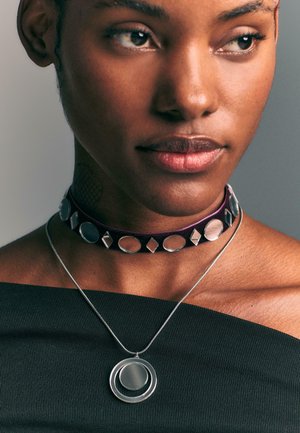 Collier ras du cou en velours violet foncé avec des accents argentés circulaires et en diamant ; pendentif en argent avec des cercles concentriques sur une fine chaîne.