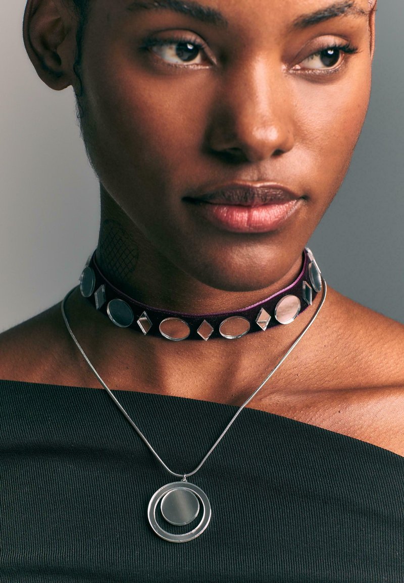 Choker-Halskette aus dunkelviolettem Samt mit kreisförmigen und diamantähnlichen silbernen Akzenten; silberner Anhänger mit konzentrischen Kreisen an einer dünnen Kette.
