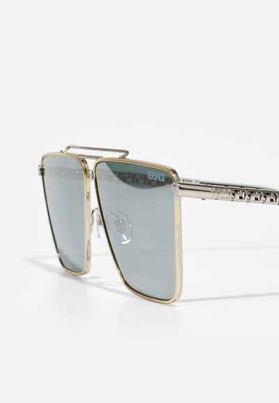 Ochelari de soare pătrați cu cadru din metal argintiu, accente aurii și lentile gri. Prezintă un pod proeminent și brațe ale templului cu un model decorativ.