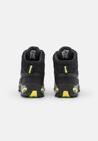 Par de zapatos de senderismo negros y amarillos vistos desde atrás, con cuellos acolchados, lengüetas y suelas rugosas sobre un fondo blanco.