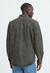 Dunkelgrüne Denim-Bluse mit geknöpften Manschetten und Kragen, die einen lockeren Schnitt und eine weiche Textur aufweist. Von hinten mit lässigen blauen Jeans gezeigt.