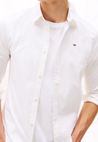 Man som bär en vit skjorta i knäppvariant över en vit t-shirt, med uppvikta ärmar och en liten logotyp på bröstet.