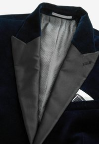 Veste de smoking en velours bleu marine avec revers crantés en satin noir, doublure intérieure grise à motifs et pochette blanche à pois noirs et blancs.