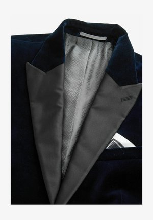 Námornícky zamatový blejzer s hrotitými klopami, šedá saténová podšívka so geometrickým vzorom a vreckový štvorcový kapesník viditeľný vo vrecku na prsiach.