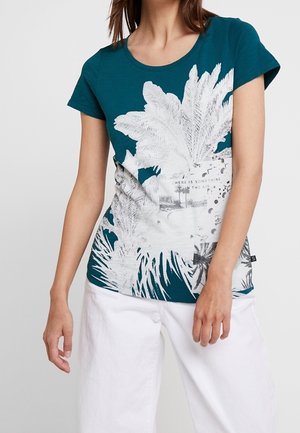 Žena v tyrkysovém tričku s krátkým rukávem s velkým bílým potiskem palmového listu a bílých širokých kalhotách, stojící před jednobarevným pozadím.