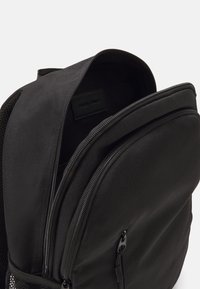 Zaino nero realizzato in tessuto testurizzato con un compartimento principale con zip e tasca frontale. Presenta una tasca laterale in rete e un'etichetta di marca all'interno.