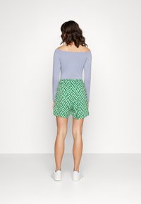 Haut bleu à manches longues dénudées, associé à un short vert à motifs avec des losanges géométriques blancs, porté avec des baskets blanches.