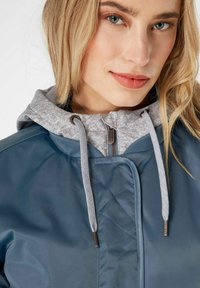 Giacca impermeabile blu con cappuccio foderato in pile grigio. Presenta un motivo a ricami sul davanti, coulisse a contrasto e chiusura con zip.