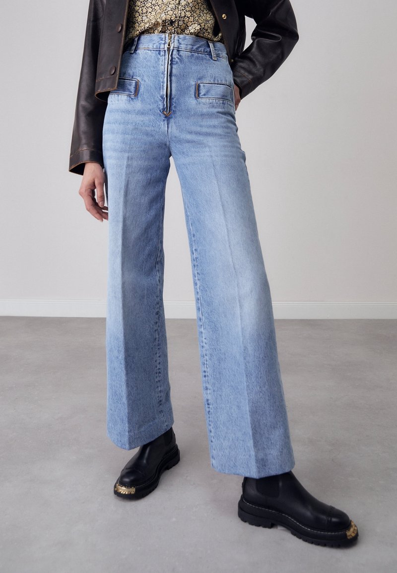 Personne portant un jean bleu clair taille haute à jambes larges, une veste en cuir noire, une chemise fleurie et des bottes noires avec un détail doré sur un sol gris.