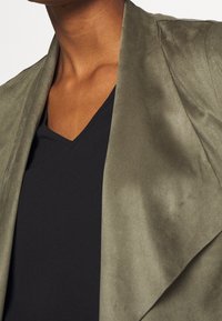 Giacca in tessuto morbido verde oliva con design drappeggiato, abbinata a un top nero a scollo a V. La texture liscia e le linee pulite sono caratteristiche notevoli.