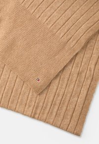 Beige textured blanket with ribbed design. One corner features a small logo tag with red, white, and blue colors. Soft, warm fabric.