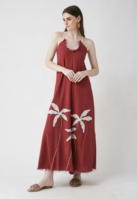 Bordeaux-maxikleid mit ausgefranster Halslinie und weißer Blattstickerei. Der Stoff ist weich, mit einer lockeren Silhouette und Seitenschlitzen. Getragen mit braunen Sandalen.