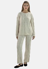 Kabelstrikket trøje og matchende bukser i off-white. Blødt, tekstureret stof med ribstrikkede manchetter og kanter. Sorte flade sko fuldender outfittet.