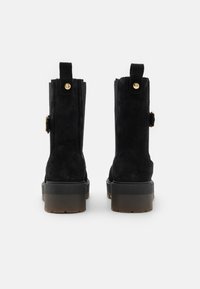 Bottines en daim noir avec une semelle épaisse, dotées d'une fermeture éclair et d'un accent doré sur le côté, ainsi qu'une languette à l'arrière pour un enfilage facile.