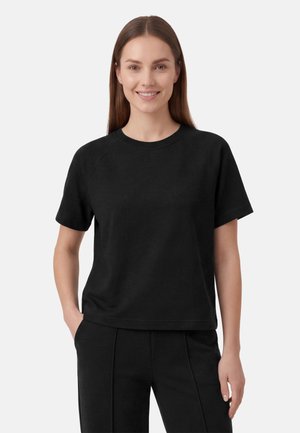 Jonge vrouw met lang, stijl haar, die een eenvoudig zwart T-shirt met korte mouwen en een zwarte broek draagt, glimlachend met één hand in haar zak.