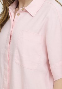 Chemise rose pâle à manches courtes avec une poche avant et deux boutons bruns près du col portée par une personne.