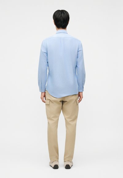 Άνδρας με πλάτη προς τα εμπρός που φοράει ανοιχτό μπλε πουκάμισο με μακριά μανίκια, μπεζ παντελόνι cargo και μπεζ παπούτσια πάνω σε απλό λευκό φόντο.