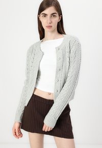 Gray knitted cardigan with a textured cable pattern, six buttons down the front, and ribbed cuffs. Worn over a white top and brown striped skirt.