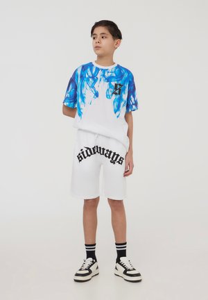 Maglietta bianca con design astratto blu e logo nero, abbinata a pantaloni corti bianchi con la scritta "sideways" in carattere nero. Sneakers in bianco e nero.