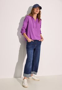 Lilla langærmet top med hætte og snore, parret med mørkeblå brede jeans og hvide sneakers med grønne accenter.
