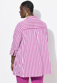 Blusa a righe rosa e bianche con maniche a tre quarti, caratterizzata da una piega sul retro e fessure laterali per facilitare il movimento.