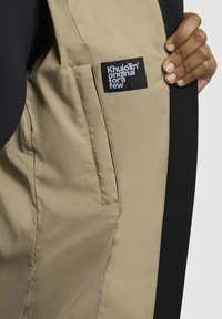 Tans kleurige buitenstof met een gladde textuur, zwarte binnenvoering en een logo-patch. Bevat een zijvak in het tan gebied.