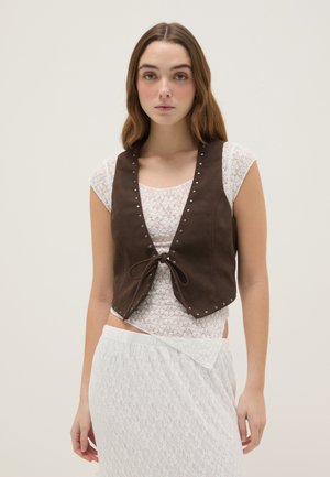 Giovane donna che indossa un gilet marrone senza maniche sopra una maglietta di pizzo bianca e una gonna in pizzo bianca abbinata, in piedi davanti a uno sfondo semplice.