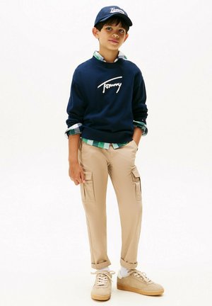Ragazzo che indossa una felpa navy di Tommy, una camicia a quadri verde, pantaloni cargo beige, sneakers beige e un cappellino navy di Tommy, in piedi con le mani nelle tasche.