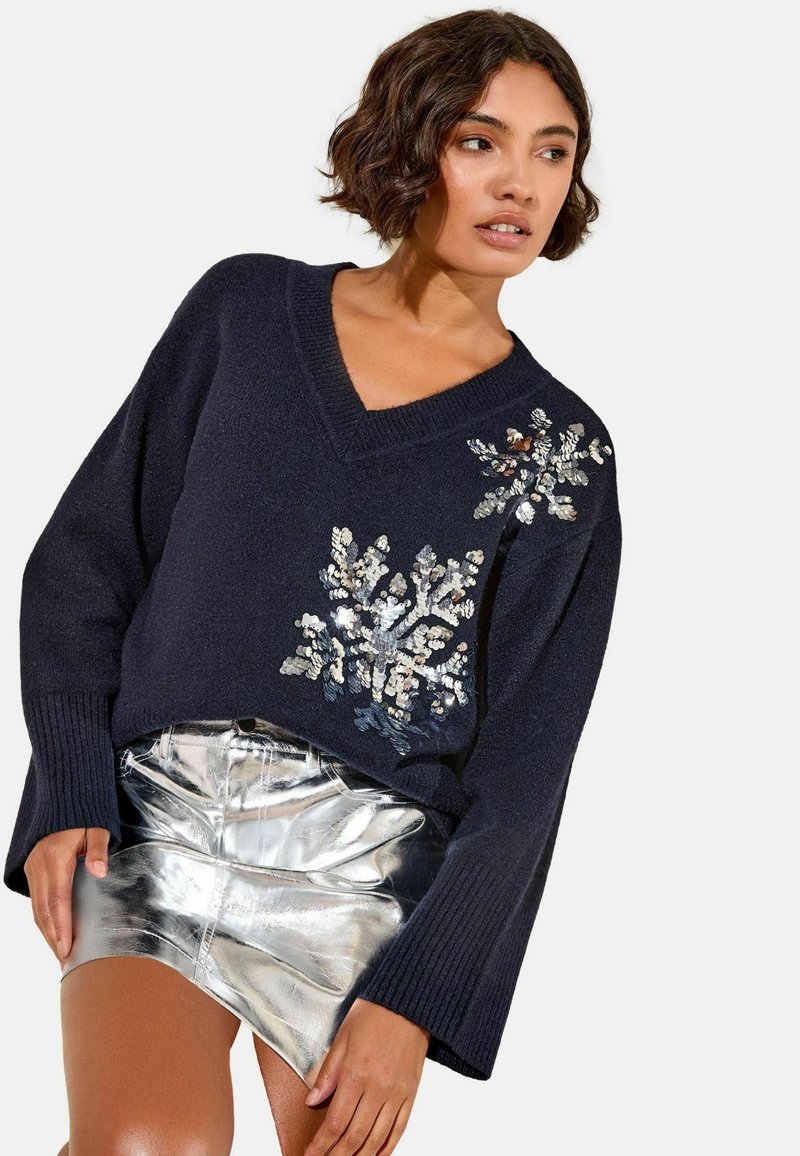 Pulover în formă de V, de culoare bleumarin, cu aplicații de paiete în design floral; asortat cu o fustă mini strălucitoare de culoare argintie. Țesătură cu tricot texturat.