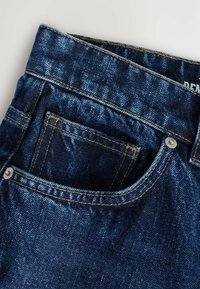 Jeans de mezclilla con un lavado oscuro, que presentan un bolsillo frontal, costuras visibles en marrón claro, un botón de metal y una etiqueta en la cinturilla.