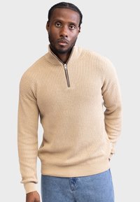 Beige genser med ribbestruktur, høy hals og glidelås halvveis. Lange ermer med noe løs passform. Enkelt design.