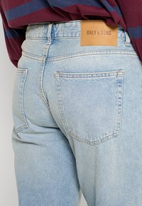 Jeans de denim azul claro con forma ajustada, que presentan bolsillos traseros y una etiqueta de cuero beige de la marca en la cintura. Textura fina y detalles de costura.