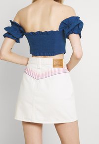 Top corto blu a spalle scoperte con maniche arricciate; abbinato a una gonna di denim bianca con un design ricamato in rosa e viola sulla vita.