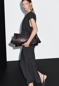 Borsa tote in pelle marrone con forma allungata e doppio manico. Il modello indossa un vestito senza maniche grigio scuro con spacchi laterali, che mette in risalto la texture liscia.