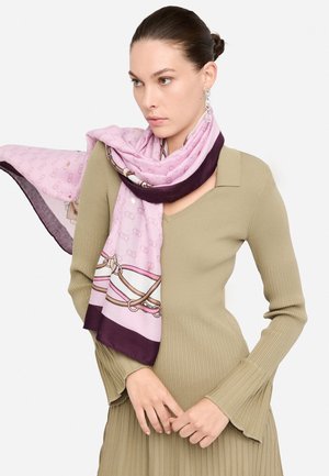 Foulard di seta in morbido rosa con un bordo viola, caratterizzato da un motivo a corde. Indossato in modo leggero su un maglione a coste verde oliva.