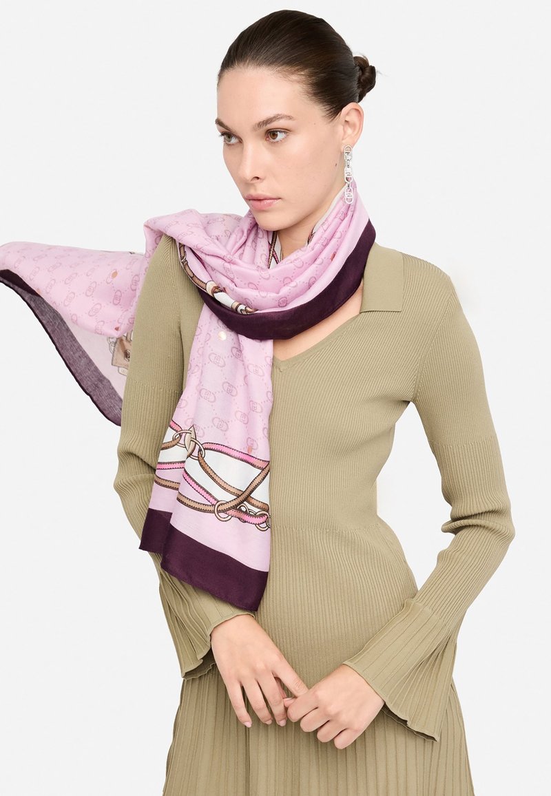 Foulard di seta in morbido rosa con un bordo viola, caratterizzato da un motivo a corde. Indossato in modo leggero su un maglione a coste verde oliva.