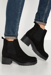 Czarne botki za kostkę z fakturowaną cholewką, elastycznymi bocznymi panelami i grubą podeszwą. Noszone z dżinsami, eksponując detal mankietu.