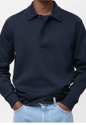 Mand iført en mørkeblå langærmet poloshirt stoppet ned i lyseblå jeans med hænderne i lommerne og et sort bælte.