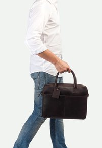 Serviette en cuir marron foncé avec deux poignées et une petite étiquette. Elle présente une texture lisse et un design simple, avec une fermeture éclair et des poches avant.