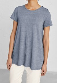 Grå kortärmad topp i mjukt material med rundad halsringning och tre dekorativa knappar på axeln. Enfärgad design.