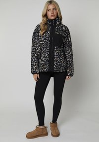 Fleecejacka med svart, beige och brun leopardmönster, hög krage, dragkedja och en bröstficka; kombineras med svarta leggings och bruna stövlar.